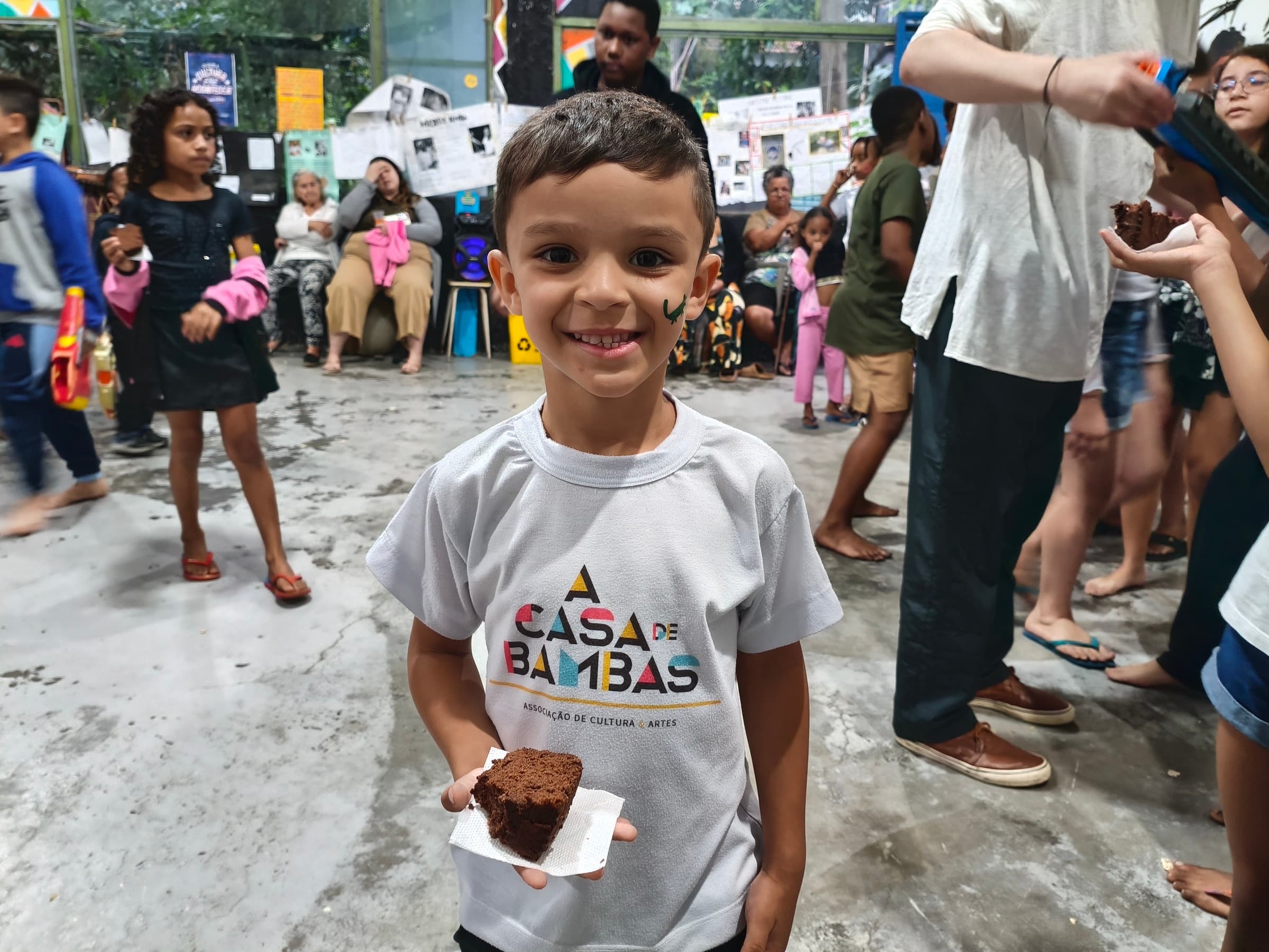 Criança sorrindo com a camiseta da Casa de Bambas, segurando um pedaço de bolo durante atividade comunitária